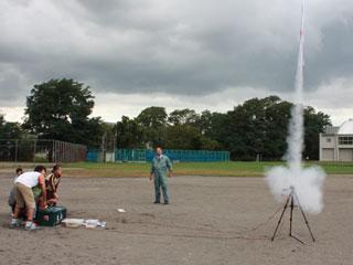 学校のグラウンドで煙を出しながら三脚から勢いよく発射する手作りロケットと、それを見守る人たちの写真
