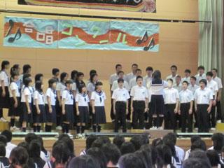 制服を着た生徒たちが壇上で大きく口をあけて歌を歌っている様子の写真