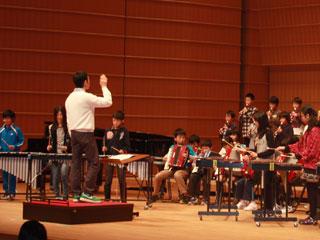 ホールの舞台で男性の指揮者に合わせて楽器を演奏する子供たちの写真