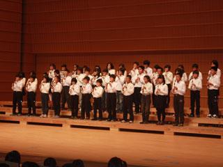 ホールの舞台で整列をしてリコーダーを吹く生徒たちの写真