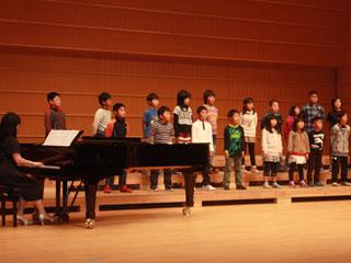 ホールの舞台で整列をして合唱する子供たちと、その横でピアノを弾く女性の写真