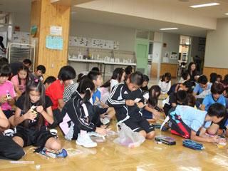 室内の床に座り折り紙を加工している大勢の児童の写真