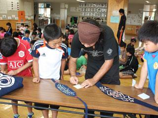 テーブルの上で児童に文様の作り方を教える、頭に布を巻いた男性の写真