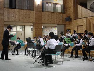 ホールで椅子に座り指揮者に合わせて楽器を演奏する生徒たちの写真