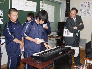 腕を組んだ男性の前で、手元のメモを見ながらキーボードを弾くジャージ姿の生徒の写真