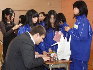 ジャージ姿の女子生徒たちの前で机に座り折り紙で鶴を折る、スーツ姿の外国人の写真