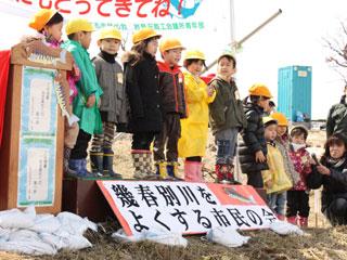 黄色い帽子をかぶった男女7名の子供たちが川沿いに設置されたステージの上で挨拶をしている様子の写真