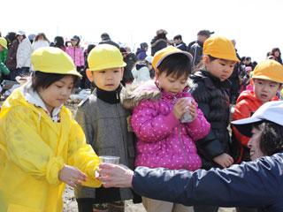 黄色い帽子をかぶった子供たちが横一列に並びサケの稚魚が入った透明なコップを順番に受け取っている写真