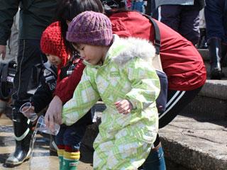 ニット帽を被った小さな子供が、寄り添う女性の手を借りてコップの中のサケの稚魚を放流している写真