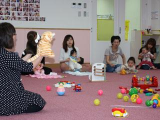 遊具が置かれた室内で、赤ちゃんとそのお母さんに手人形を披露する女性の写真