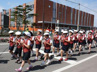 3列に並んだ制服を着た小学生たちが、リコーダーを吹きながら車道を行進する写真