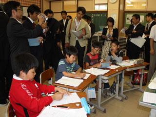 教室内で大勢の人が机に向って勉強する小学生の姿を見ている写真