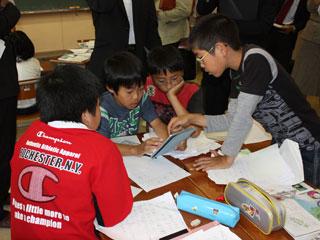 教室内の机の上で、タブレット端末を相談しながら操作する小学生たちの写真