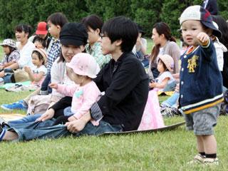 芝生にレジャーシートを敷いて寛いでいる人たちと、その近くに立っている帽子をかぶった子供の写真
