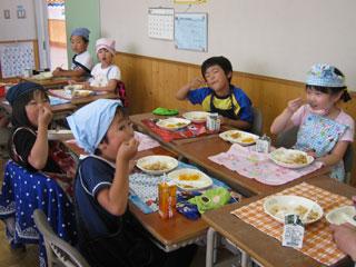 教室の机に向かい、美味しそうに給食を食べるエプロン姿の生徒たちの写真