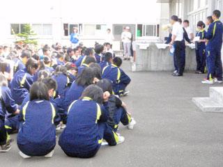 校舎の前でしゃがんで話を聞いている、大勢のジャージ姿の生徒たちの写真