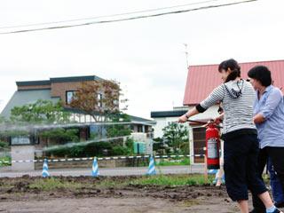 民家の近くの屋外で、消火器を使って共同で消火体験をする女性の写真