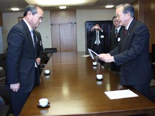 市長応接室の大きな机で男性2名づつが向かい合わせで並び、右の男性が目録を左の男性に渡そうとしている写真