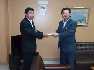 教育長室で、黒いスーツ姿の男性と紺色のスーツ姿の男性が書類を手渡している様子の写真