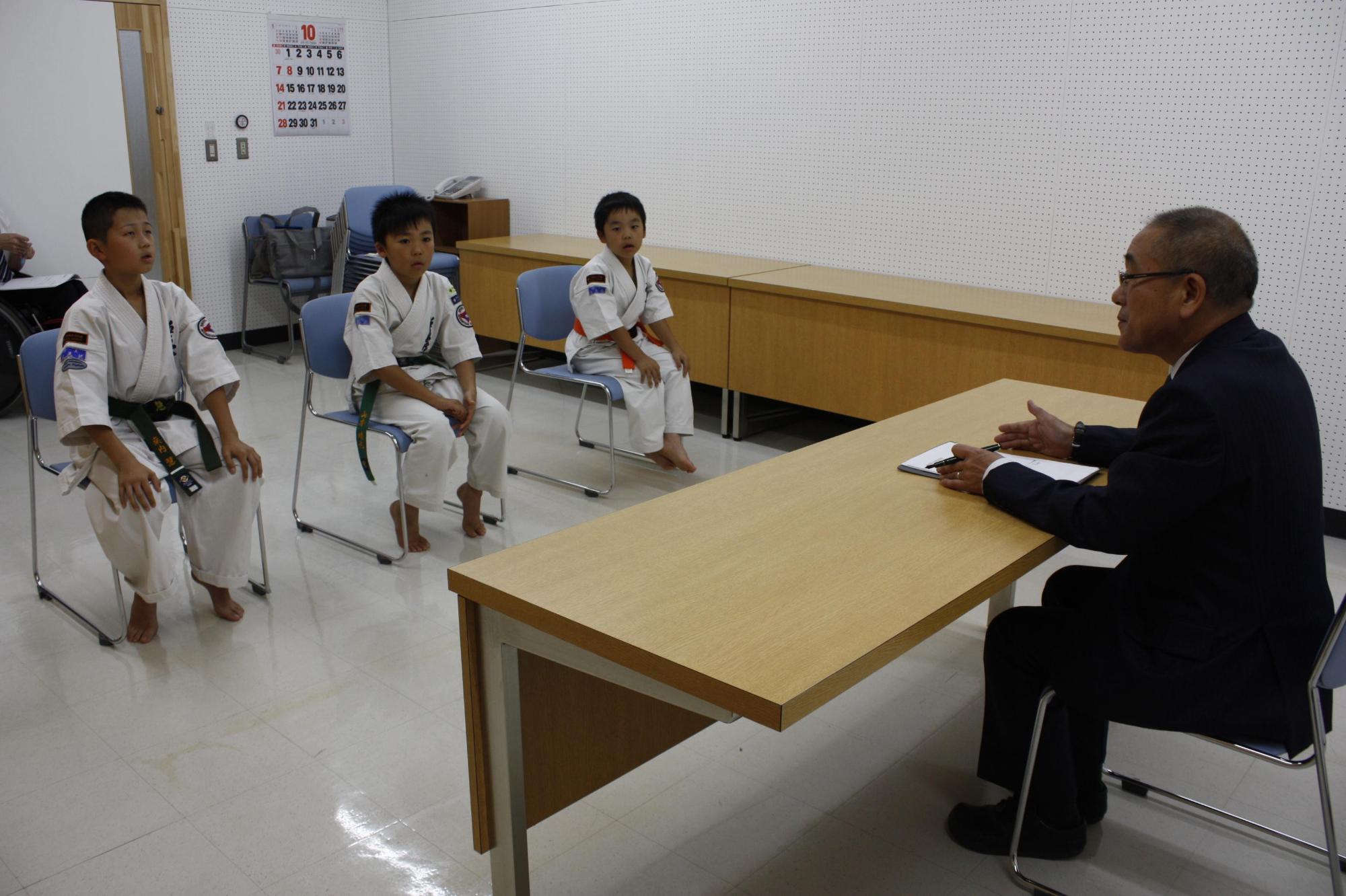 空手道着のまま教育長と向かい合って椅子に座り談笑する小学生3人の写真