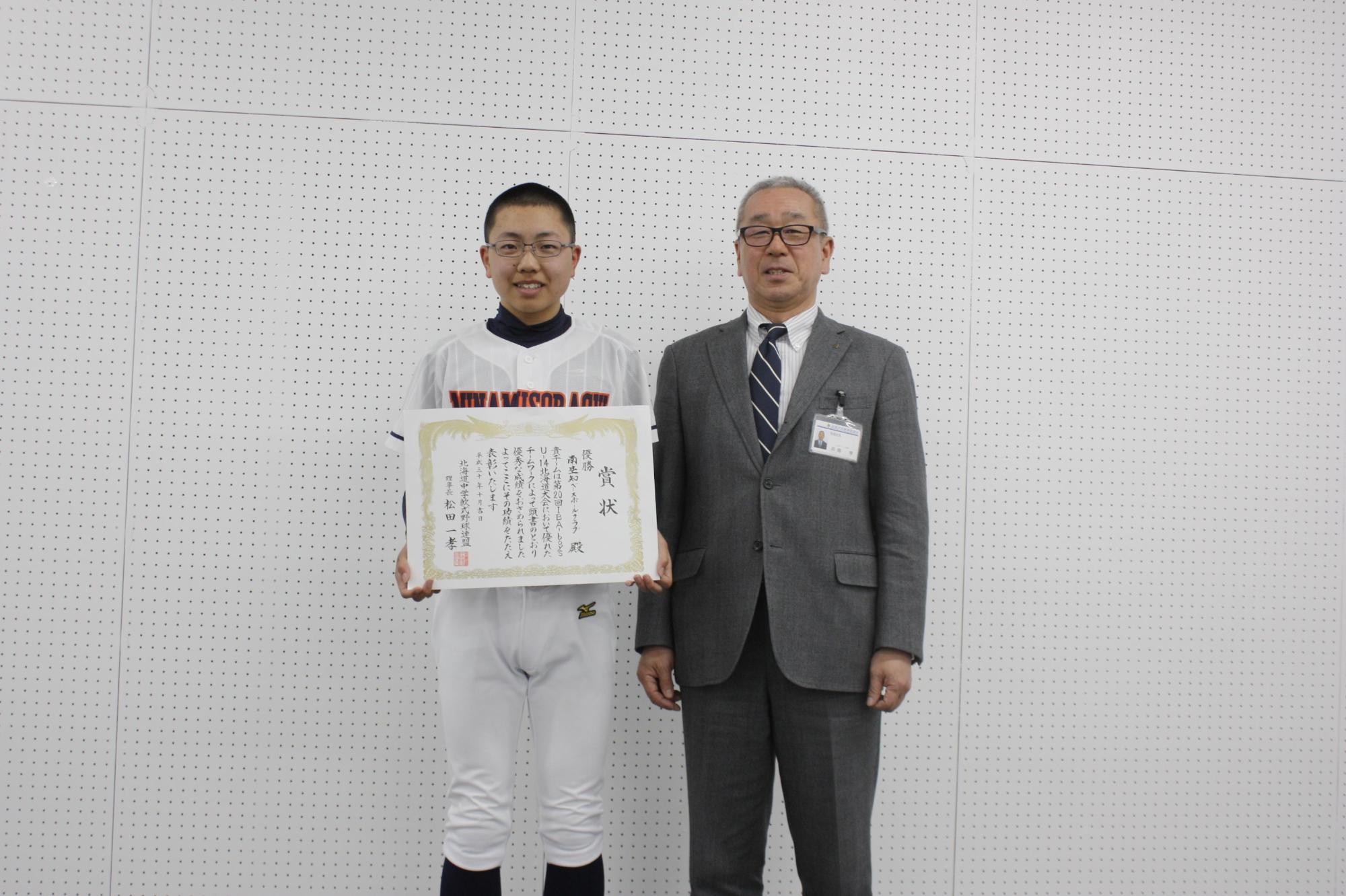 教育長の横でユニフォームを着た学生が賞状を持っている写真