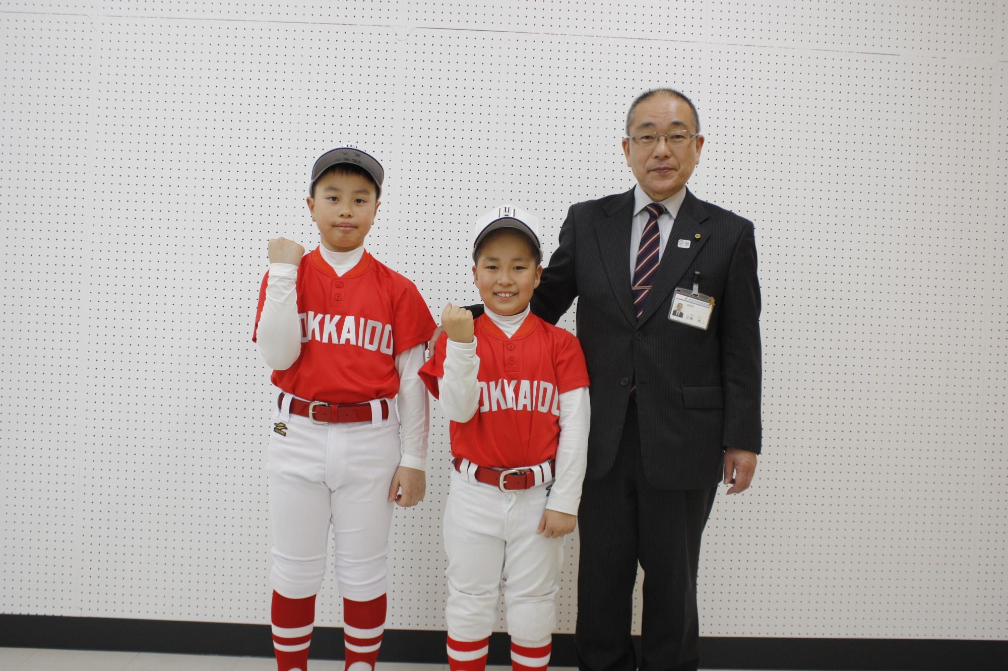 教育長の横でユニフォームを着た児童たちがガッツポーズをしている写真