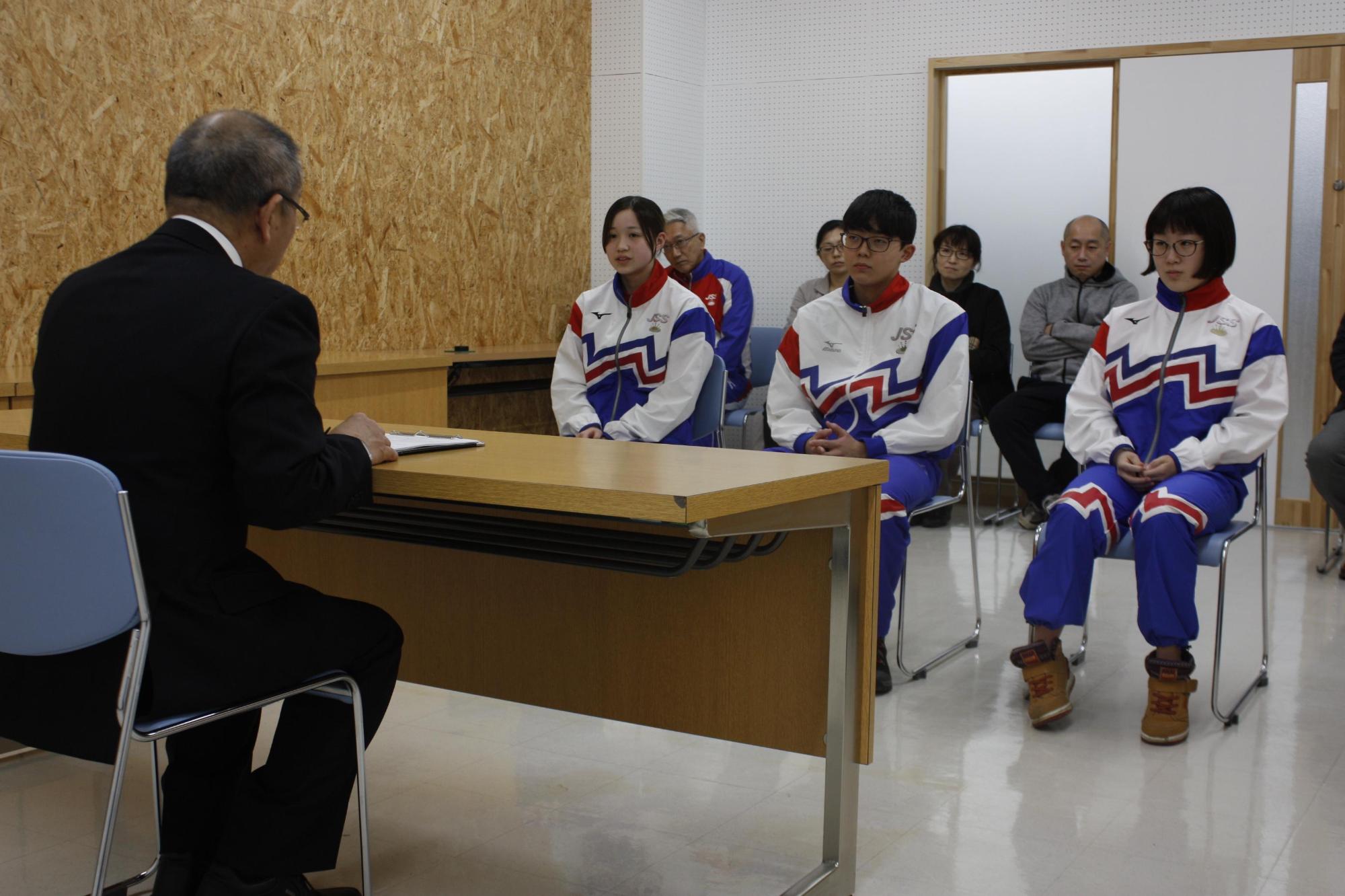 教育長の前に座っている青と白のジャージをを着た学生たちの写真