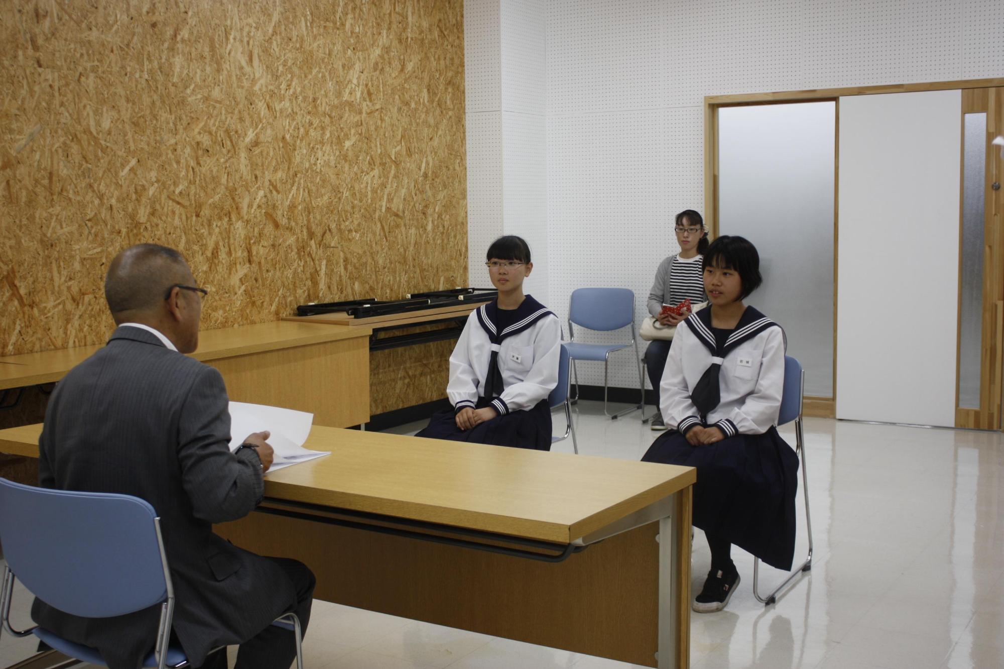 スーツを着た男性と机を挟んで、2人の白い制服を着た女子生徒と奥に一人の女性が椅子に座っている写真