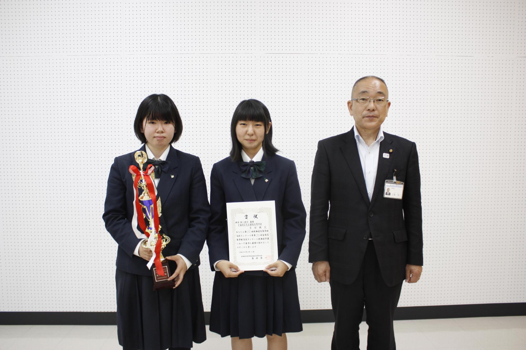 右からスーツを着た男性と、賞状を持った女子生徒と、トロフィーを持った女子生徒が並んでいる写真