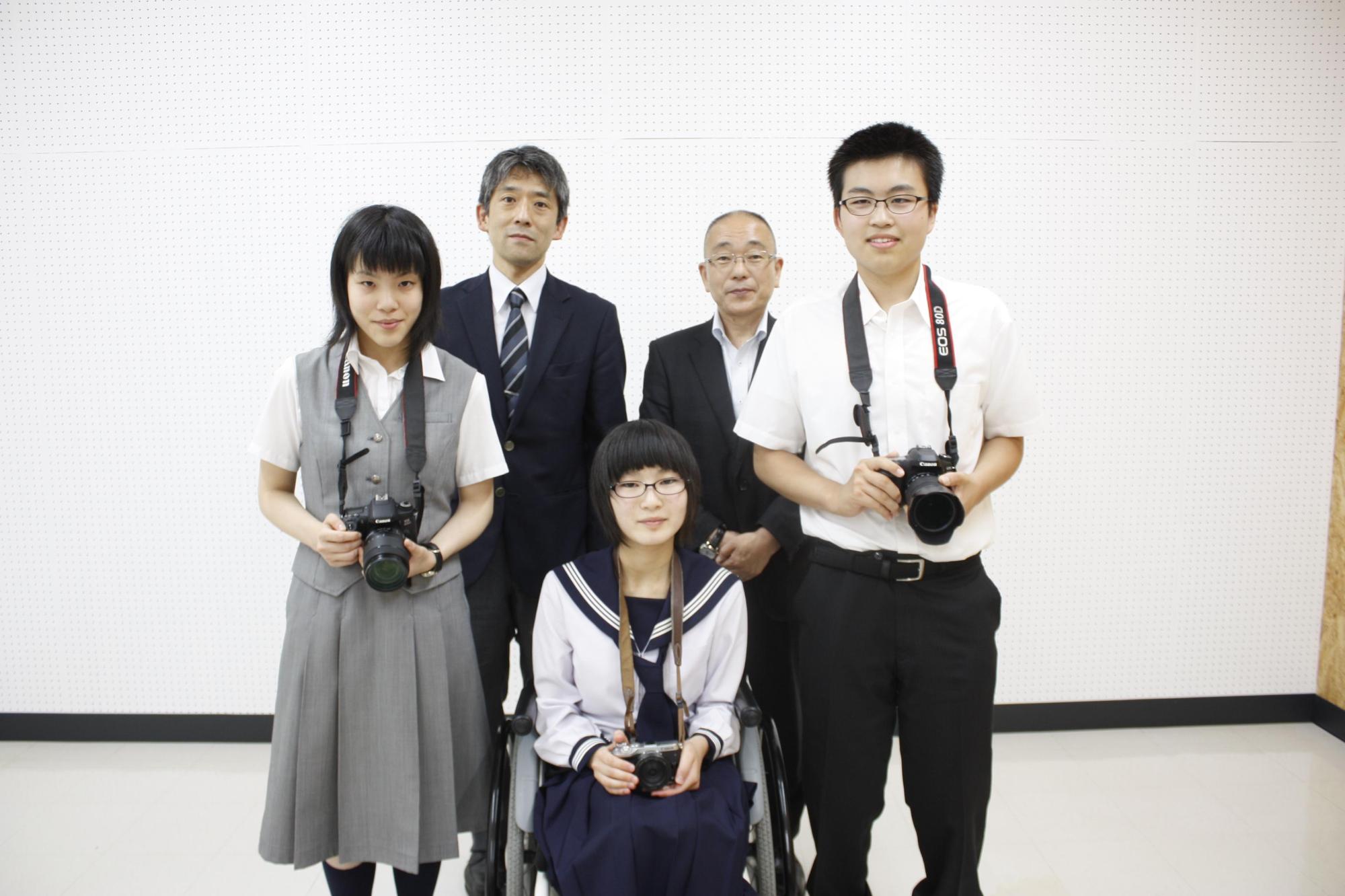 右から、カメラを首にかけて立っている男子生徒、車椅子に座っている女子生徒、カメラを首にかけて立っている女子生徒が並んでおり、その後ろのスーツを着た男性2人が並んでいる写真