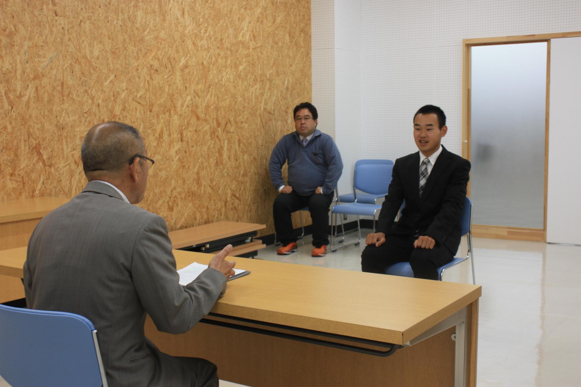 スーツを着た男性と机を挟んで、黒いスーツを着た男子生徒と、奥に1人の男性が椅子に座っている写真