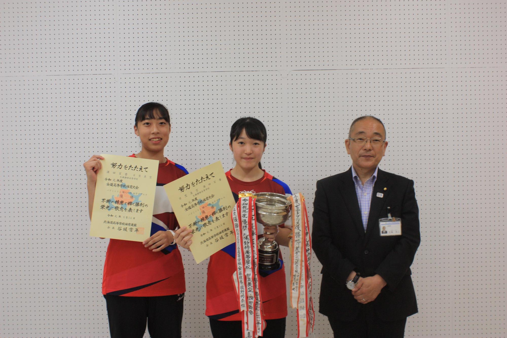 右から、スーツを着た男性と、賞状とトロフィーを持った女子生徒と、賞状を持った女子生徒が並んでいる写真