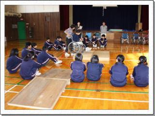 ボランティアに参加する中学生が体育館で車椅子で段差を登るお手本を見ている様子の写真