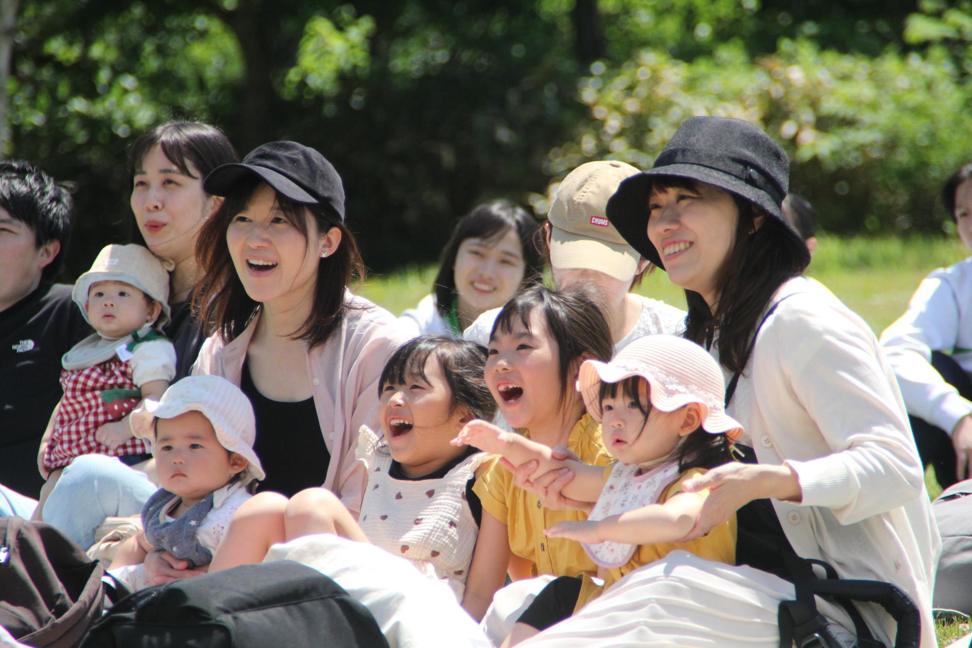 緑が生い茂る公園に座りながら、たくさんの親子が楽しい層に笑顔で写る青空広場の写真