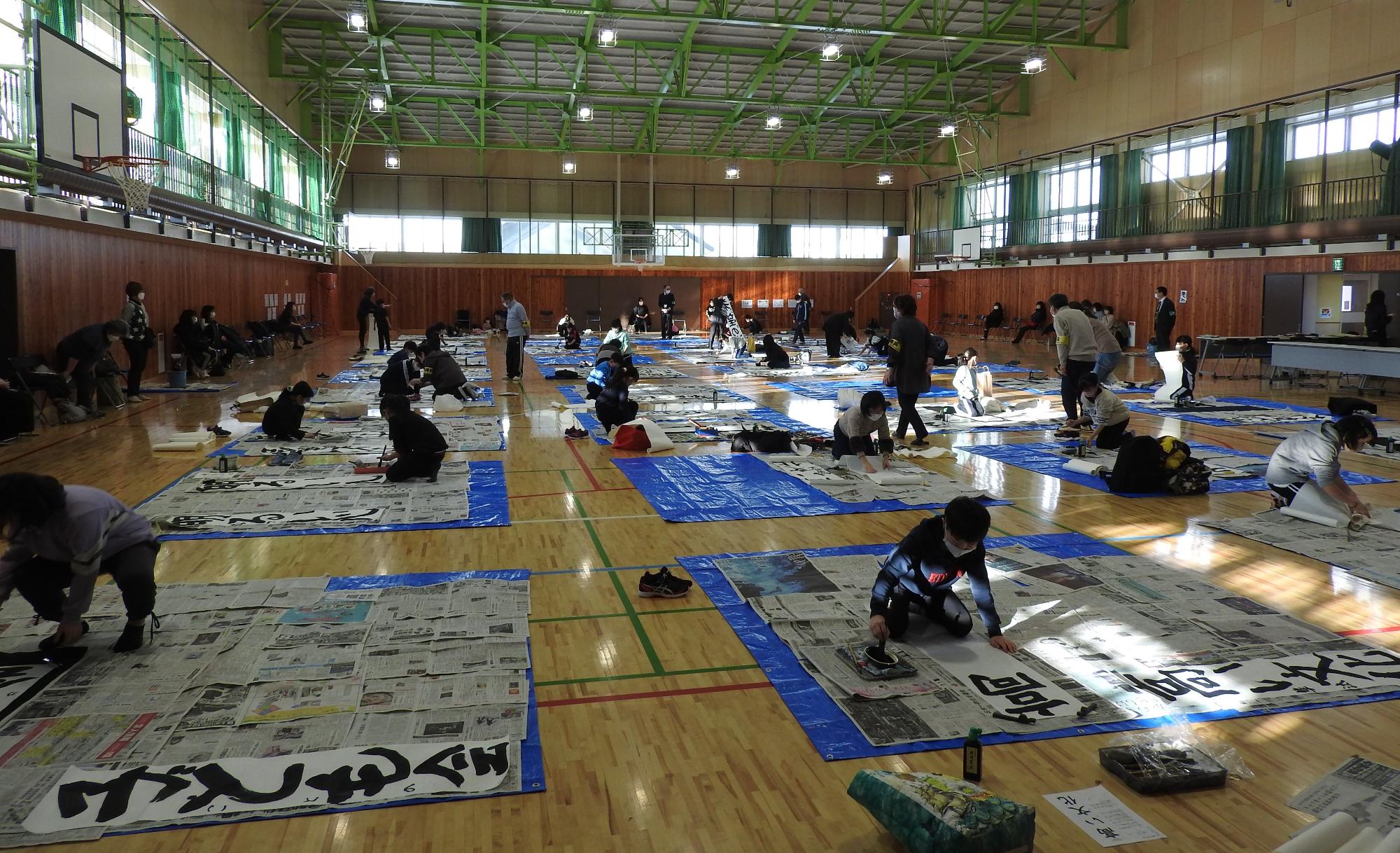 体育館の床に子どもたちが並び、半紙を広げ書初めをしている、令和4年度子ども書初め大会の写真