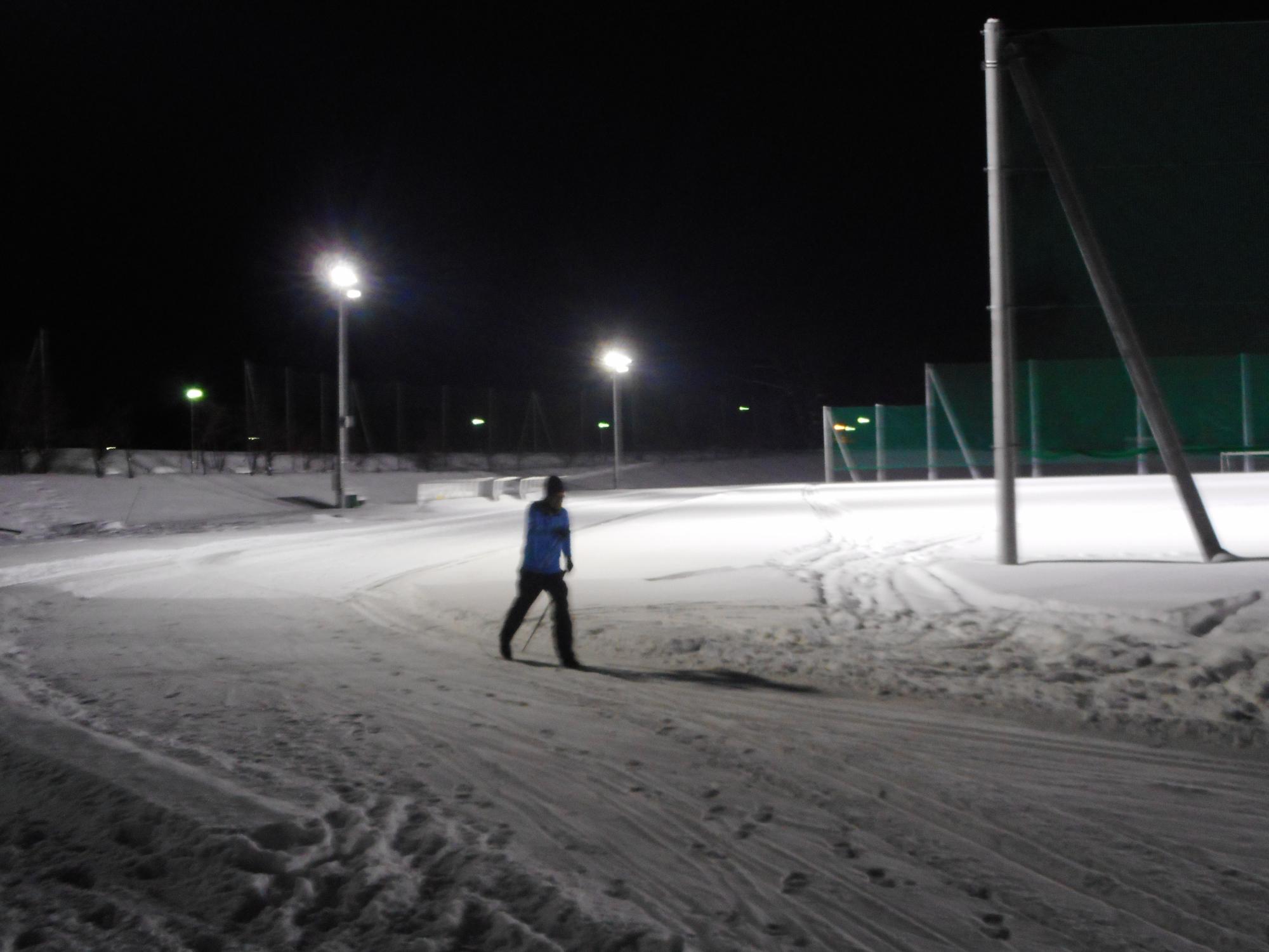 真っ暗闇の下、辺り一面雪に覆われたグラウンドを歩いている1人の人を照らす照明設備の写真
