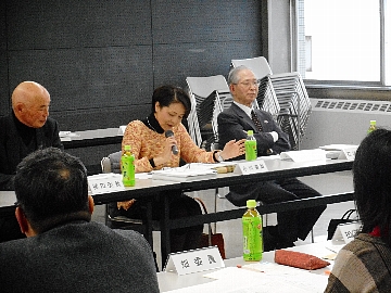 机の前に置かれた椅子に座りながら会議に参加する人々の中で、オレンジの服を着た女性がマイクを持ち話している写真