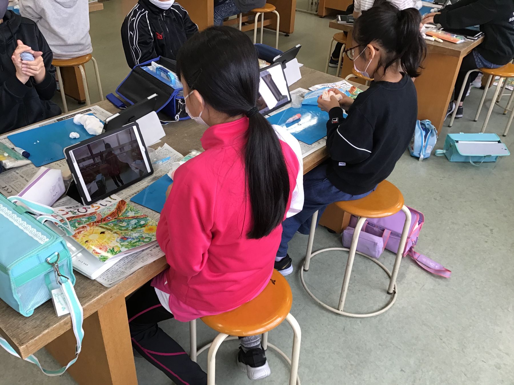 教室の机に向かい、タブレット端末を見ながら粘土を練る女の子たちの写真