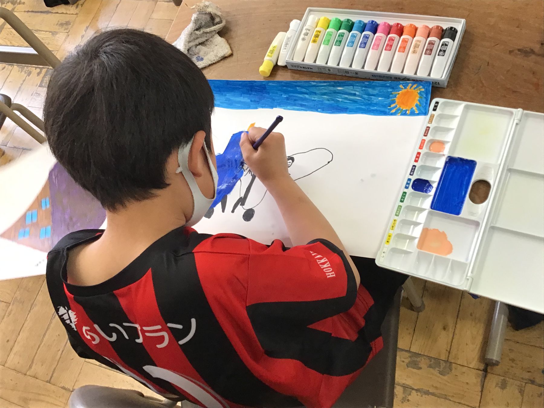 教室の机に向かい、白い画用紙に青い絵の具で絵を描く男の子の写真