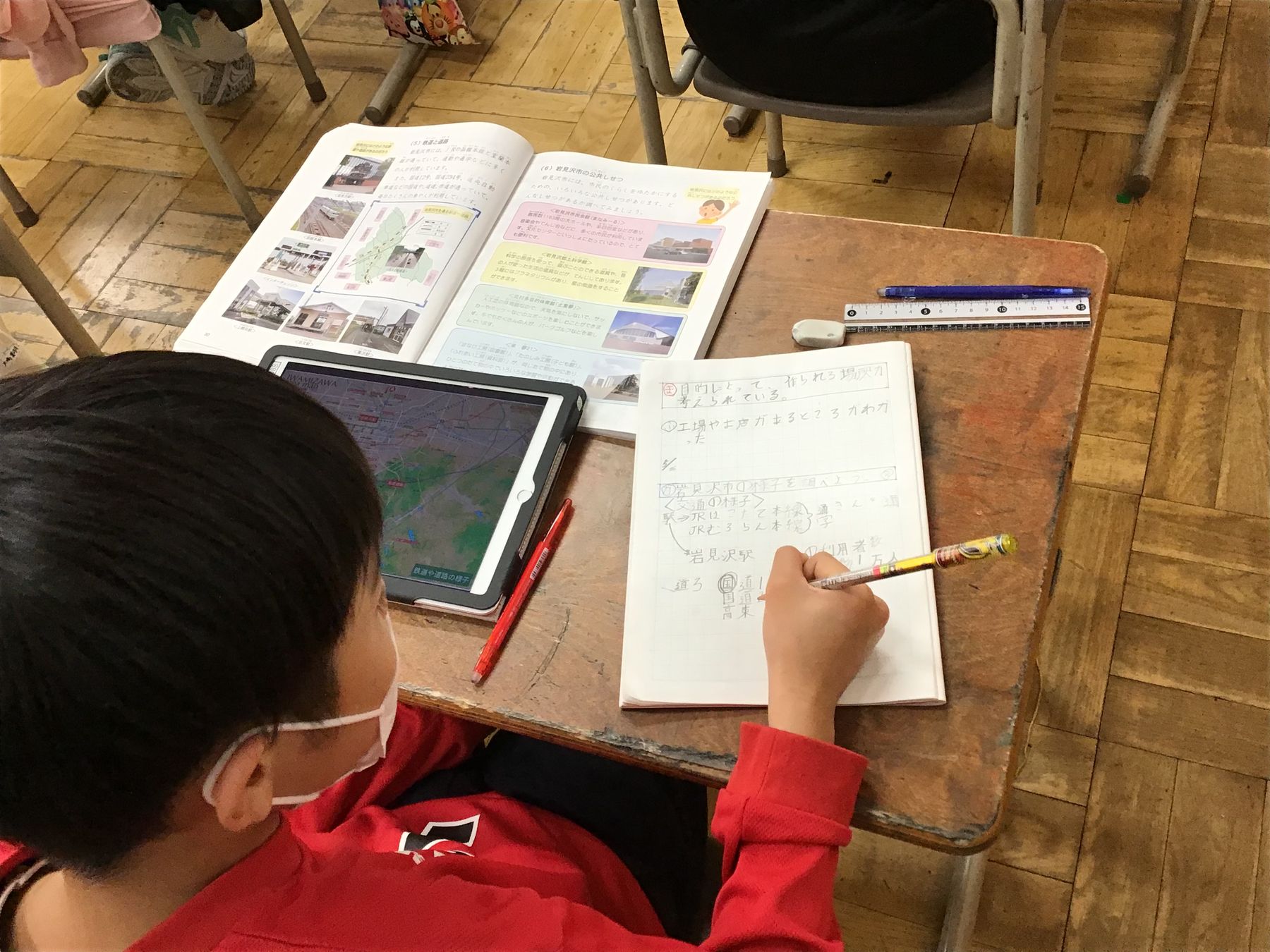 教室の机に向かいタブレット端末と教科書を見ながら、ノートに文字を書き込む男の子の写真