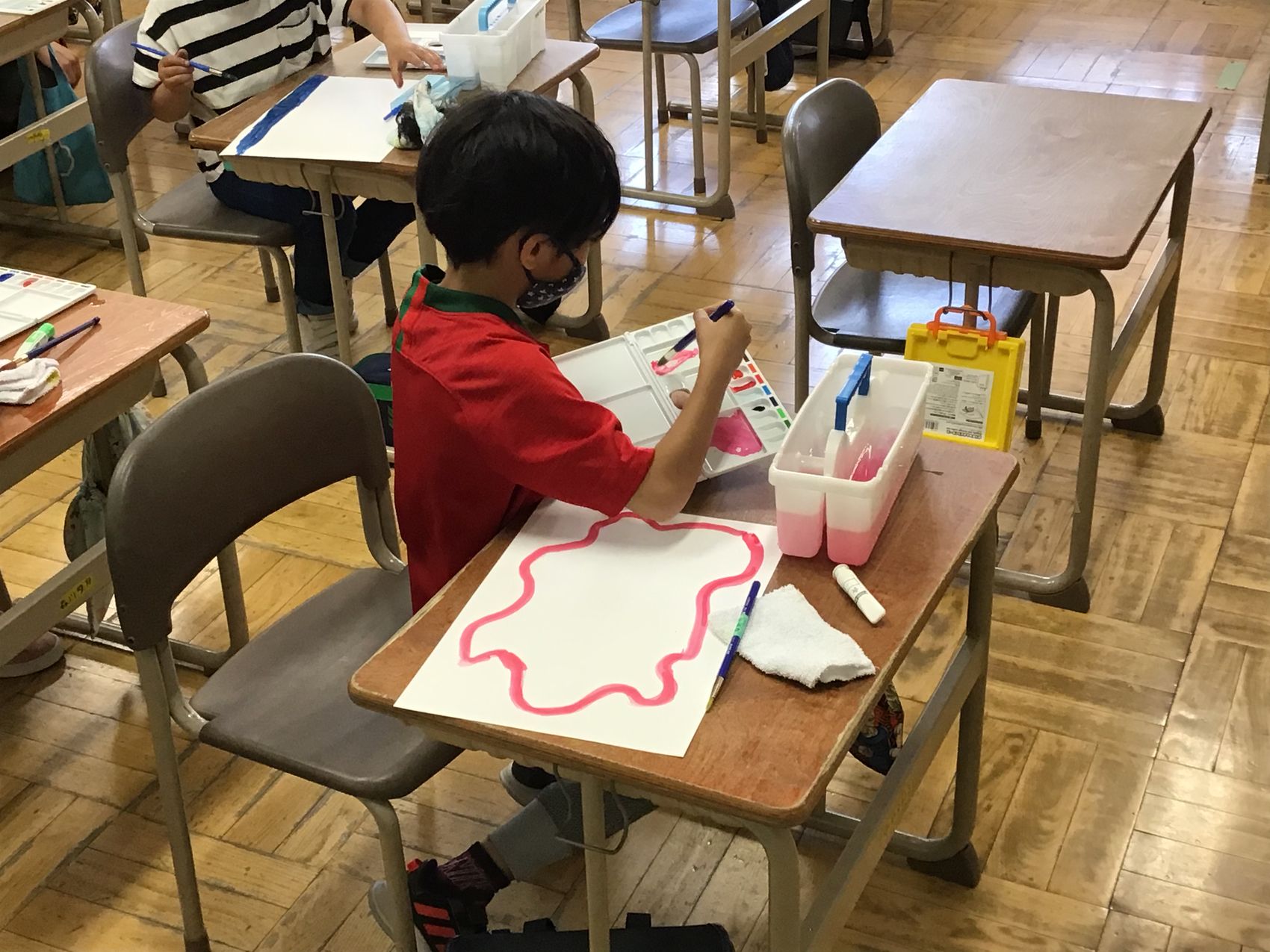 教室の机に向かい、白い紙にピンク色の絵の具で絵を描く男の子の写真