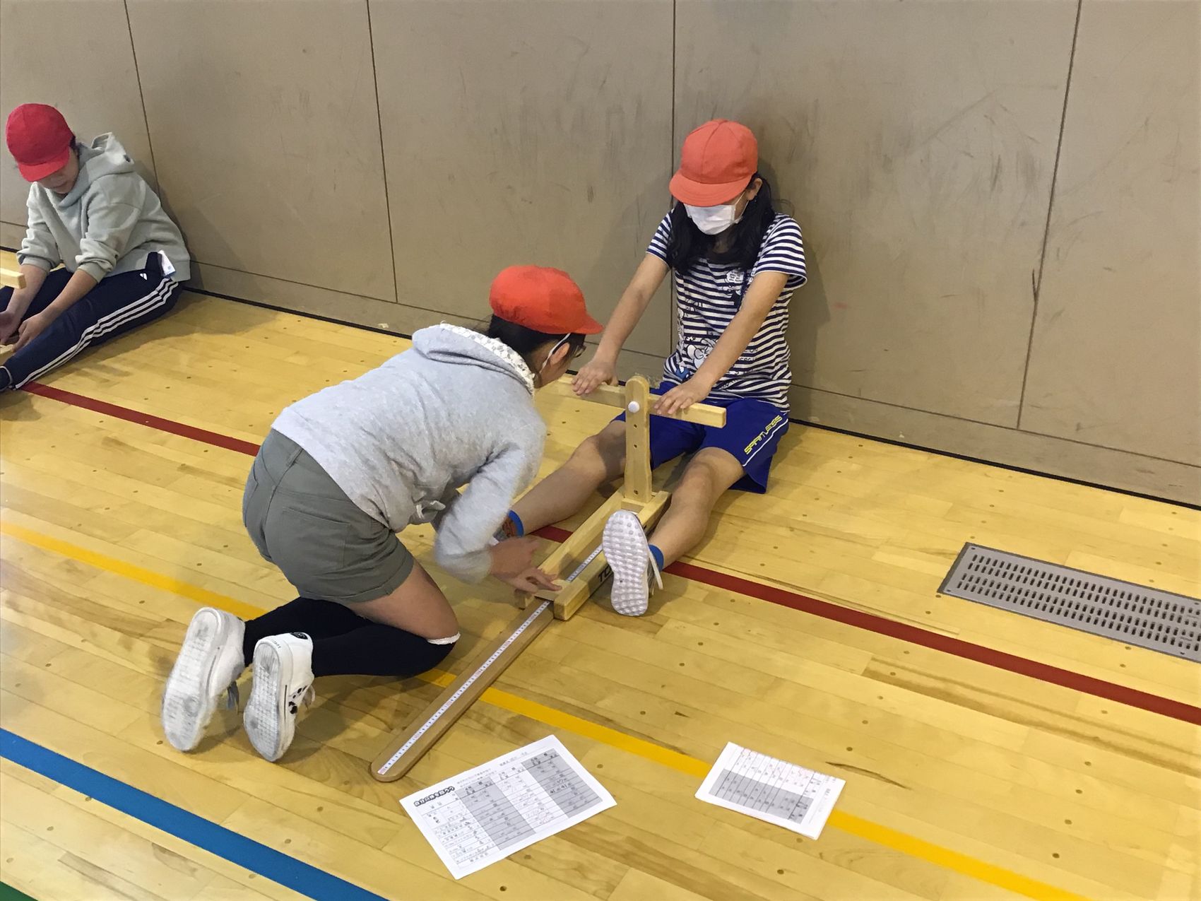体育館の床に座り木製の器具を使って体力測定をしている、赤い帽子をかぶっている2人の女の子の写真