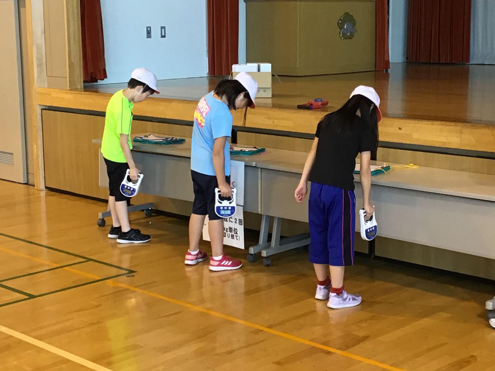 体育館の壇上の前で横一列になり、握力測定をしている白い帽子をかぶった3人の子供の写真