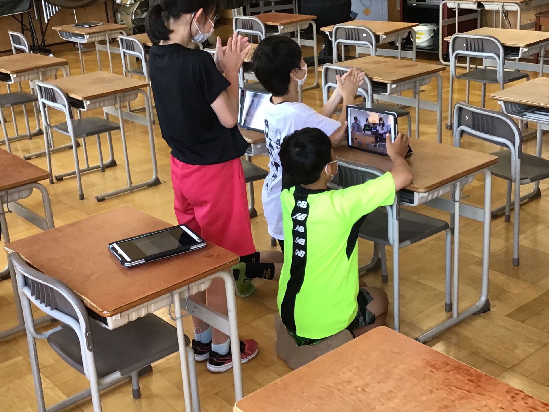 教室の机の前でタブレット端末を見たり、手を叩いている3人の子供の写真