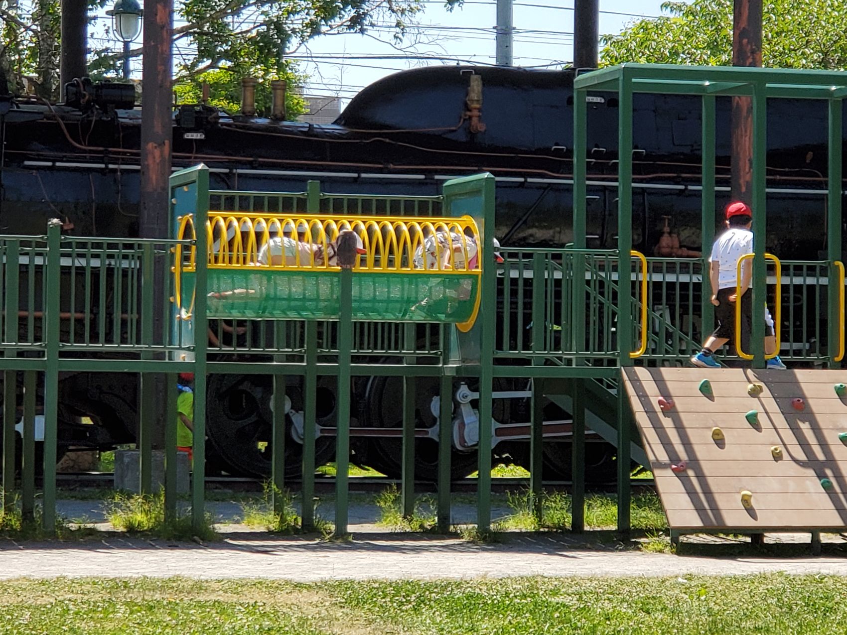 公園に設置された機関車の前にある遊具で遊ぶ、赤や白の帽子をかぶった体操着姿の子供たちの写真