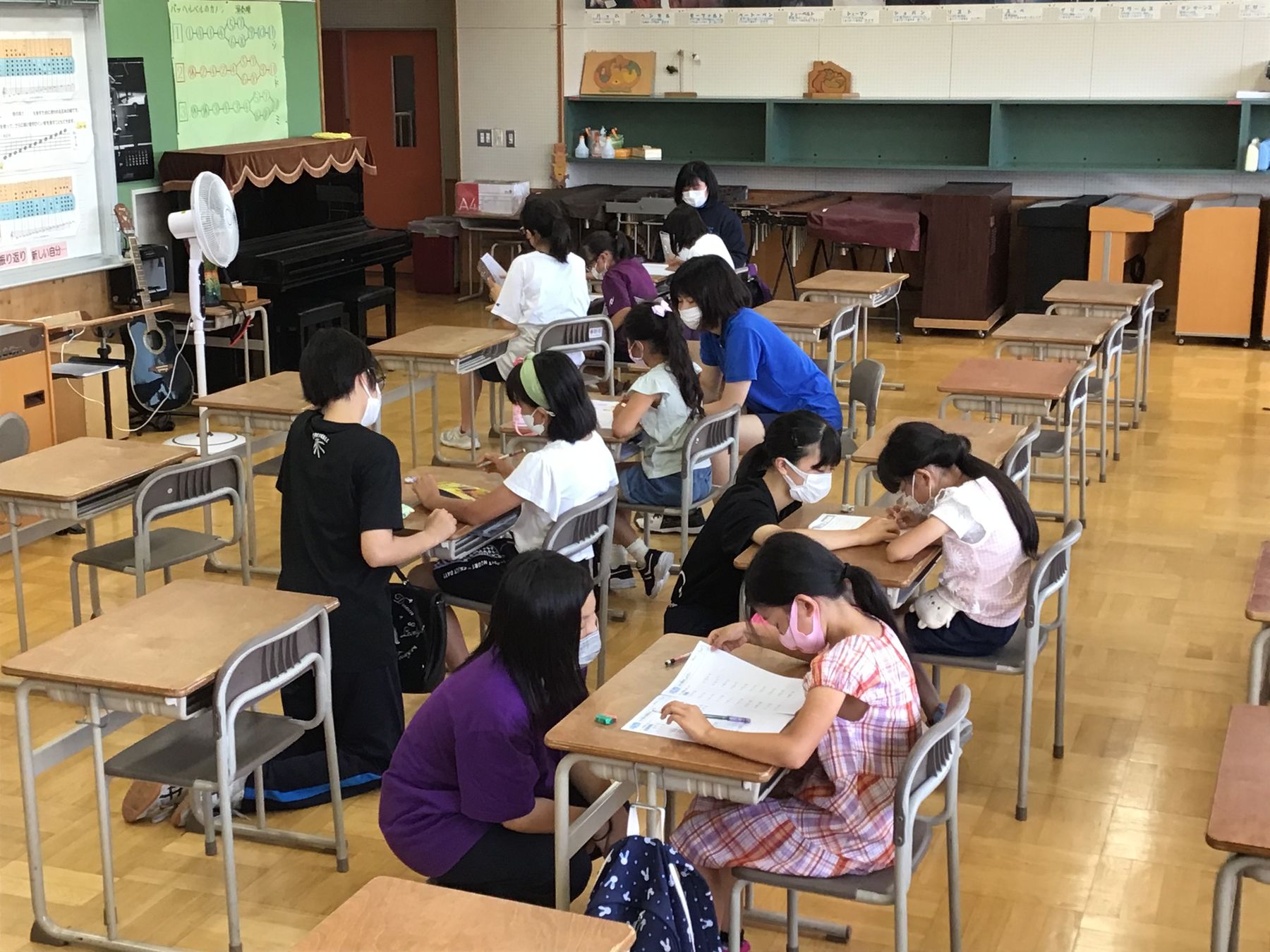 扇風機がある教室の机に向かい宿題に取り組む小学生と、目線の高さを合わせて指導する中学生の写真