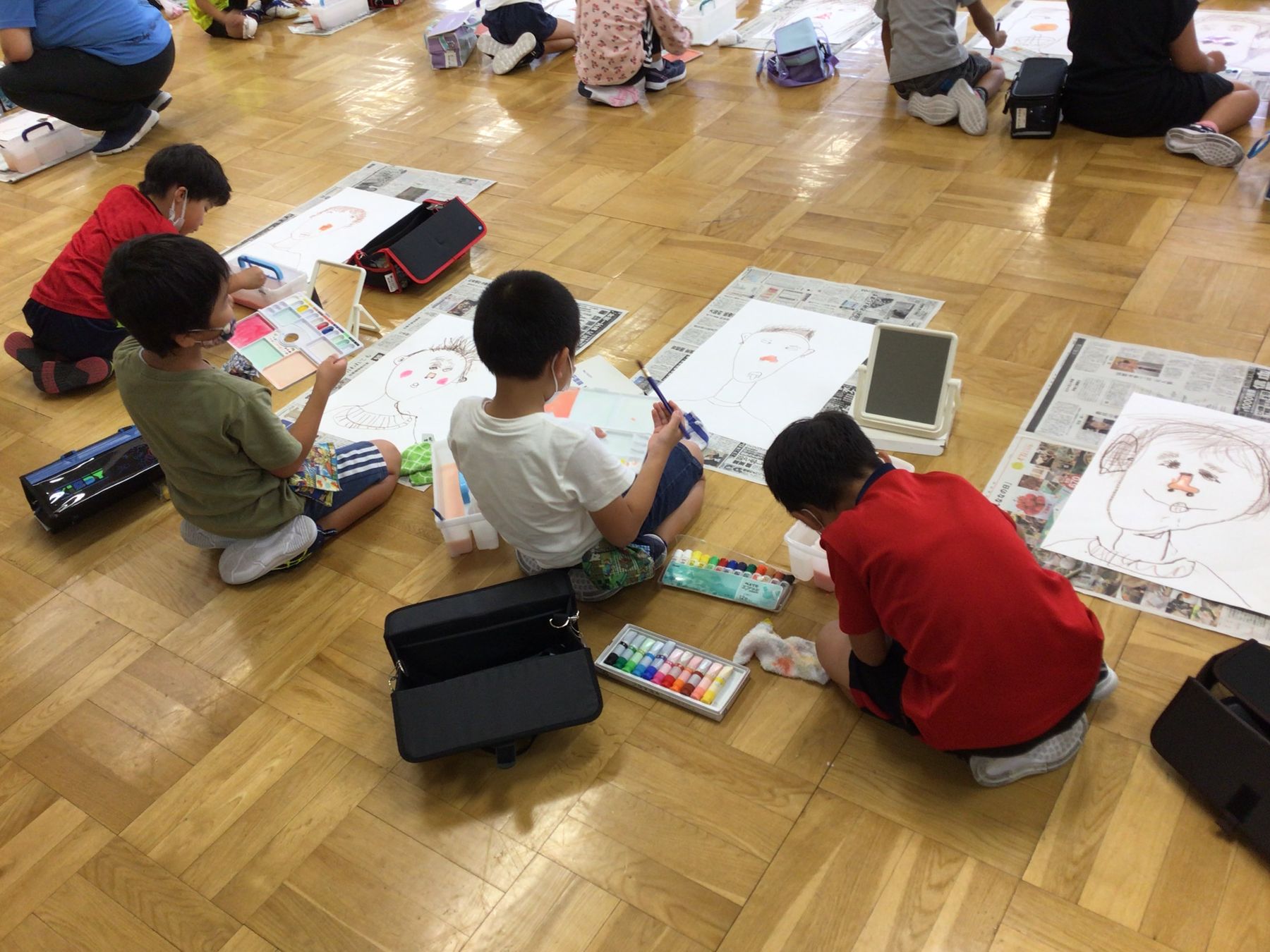 正座をして室内の床に新聞紙を敷き、カラフルな絵の具を使い白い紙に自分の顔を描いている子供たちの写真