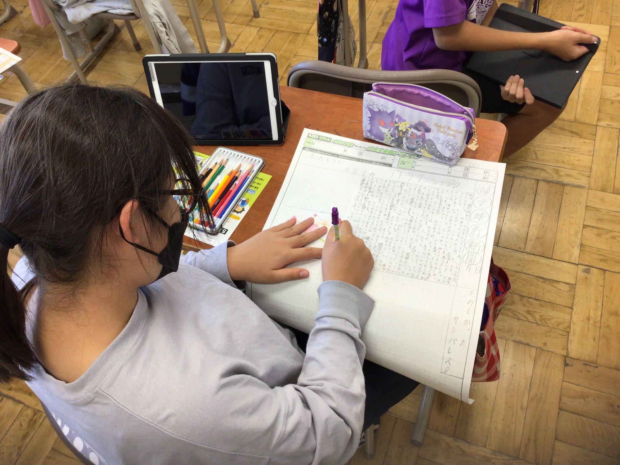 教室の机に向かい、色鉛筆を使って新聞作りをしている女の子の写真