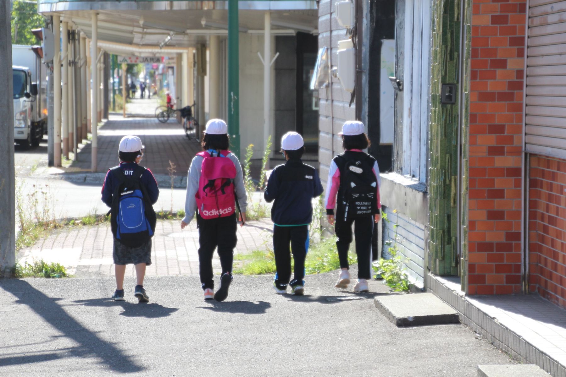 町中の歩道を横一列になって歩く、白い帽子をかぶりリュックを背負ったジャージ姿の4人の子供の写真