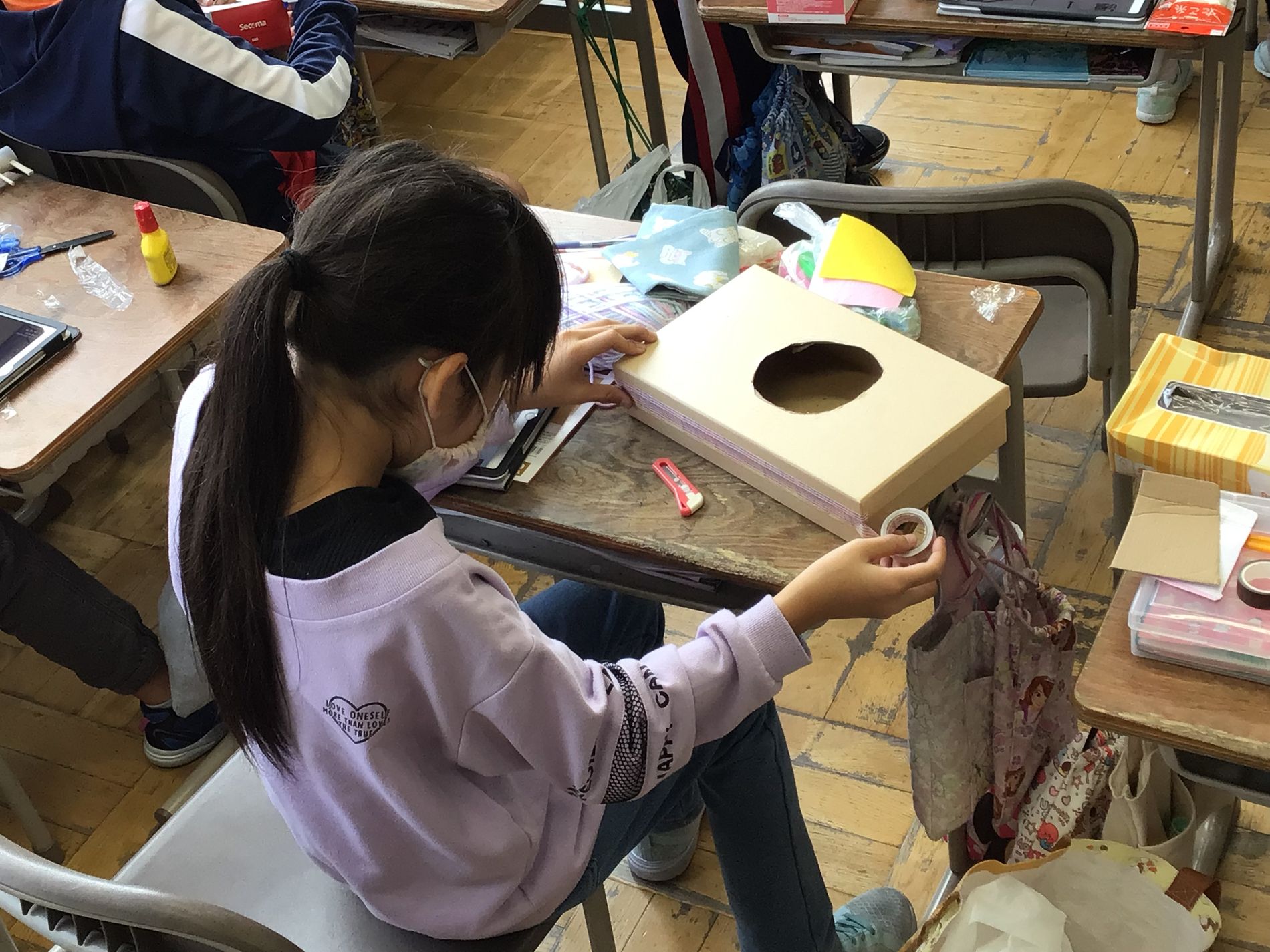 教室の机に向かい、箱型の段ボールに穴を開けたりテープを貼って加工している女の子の写真